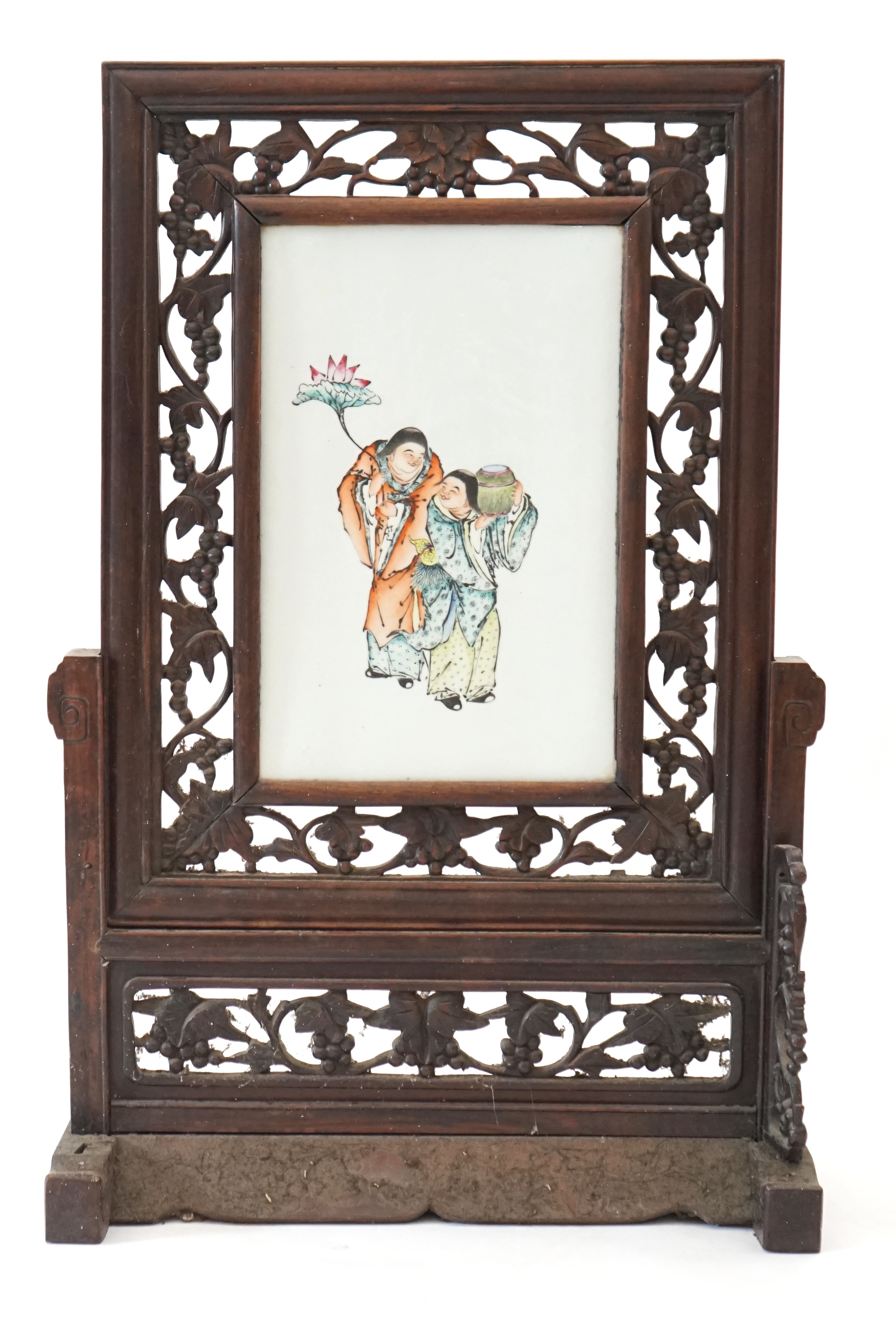 A Chinese enamelled porcelain and hardwood table screen, Republic period, painted with the HeHe Erxian, holding a sprig of lotus and a box and cover, the plaque 37.5cm x 24cm, the wood stand and frame carved with vines,